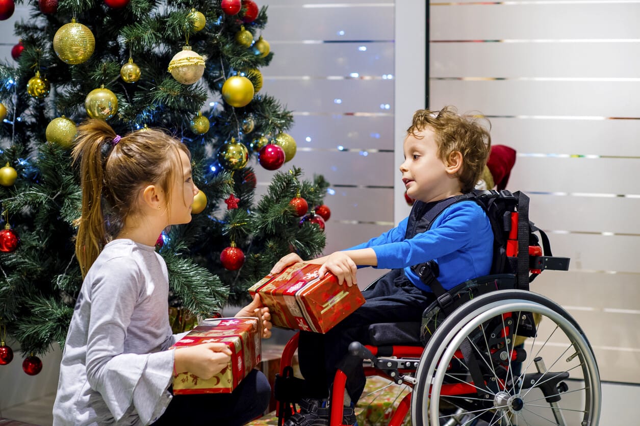 Two children exchange gifts in front of a decorated Christmas tree. One child is in a wheelchair.